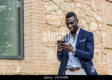 African American businessman walking with smartphone, smiling. Young professional in elegant suit sending text message, with space for text in the ima Stock Photo