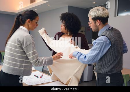 Two Women and a Man, are standing around a table, one of them holding and analyzing a piece of fabric. Scene is collaborative and focused, as the grou Stock Photo
