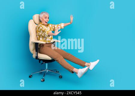 Full length photo of lovely young lady sit armchair ride have fun dressed stylish striped garment isolated on yellow color background Stock Photo