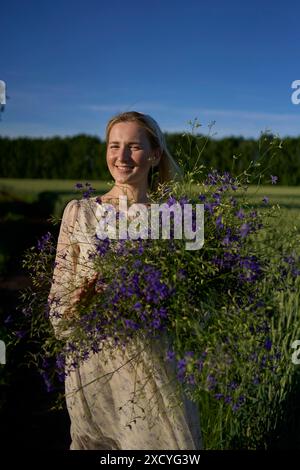 a portrait of a young blonde woman in a pastel chiffon dress walking with a giant bouquet of wild flowers in a field Stock Photo