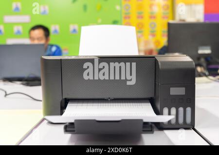 A black printer is open and has a white sheet of paper inside. The printer is on a desk in front of a computer Stock Photo