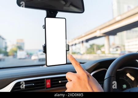 Close-up smartphone with white mockup on screen, background of car steering wheel. A person is pointing at a phone mounted on the dashboard of a car. Stock Photo