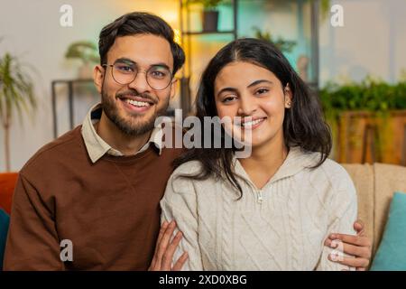 Loving young diverse couple looking at camera and smiling sitting arm around relaxing on sofa. Happy Hispanic family spending leisure time together on couch looking at camera in living room at home. Stock Photo