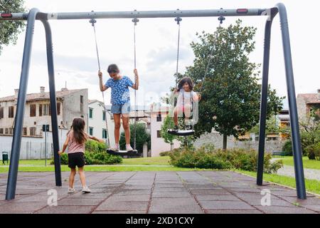 three sisters on children playground playing with swing Stock Photo