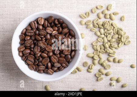 Roasted coffee beans in a white bowl on linen fabric. On the right unroasted green Arabica coffee beans, seeds of berries from Coffea arabica. Stock Photo