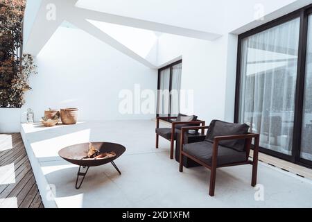 A modern outdoor patio with minimalist design featuring two wooden chairs with dark cushions, a metal fire pit, and potted plants against white walls. Stock Photo