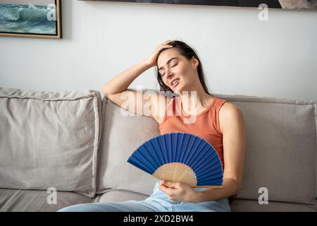Glad woman successfully coping with abnormal heat, blowing with paper fan, sitting on couch. Stock Photo