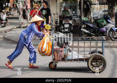 Vietnamese old woman pushing a cart with garbage bags. Hoi an, Vietnam. A woman wearing traditional Vietnamese clothing pushing metal trolley. Travel Stock Photo