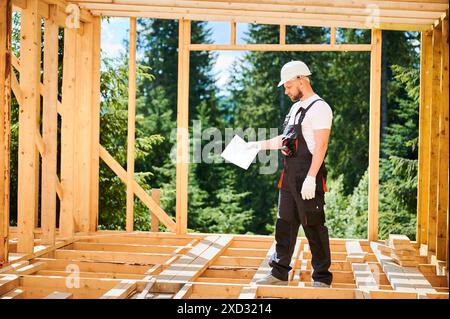 Supervisor overseeing construction of wooden two-story house located near woods. Bearded man dressed in jumpsuit and protective helmet is studying the construction plan. Stock Photo