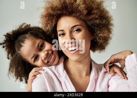 A joyful African American mother and daughter, dressed in pajamas, are striking a pose in front of a grey background. Stock Photo