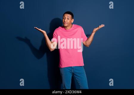 Photo of young funny guy in pink stylish t shirt confused shrug shoulders having no idea how solve it isolated on dark blue color background Stock Photo