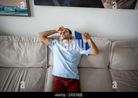Exhausted unhappy man blowing with paper fan, suffering from stuffiness heat, lying on sofa. Stock Photo