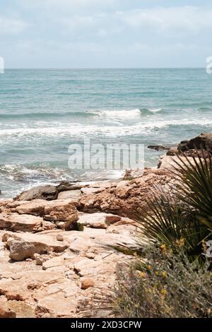 A serene view of a rocky beach with gentle waves lapping against the shore under a clear blue sky The landscape features rugged rocks in the foregroun Stock Photo