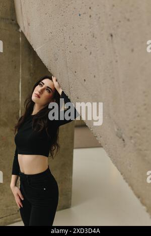 A young woman with dark hair stands near a concrete wall, looking up thoughtfully Stock Photo