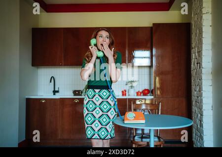 A redhead woman in a colorful 1950s-style kitchen appears shocked while talking on a classic green rotary phone. She's wearing a green top and a patte Stock Photo