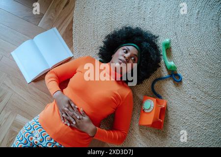 From above black African American woman dressed in a bright orange sweater and patterned skirt, laying on the floor with a vintage green telephone bes Stock Photo