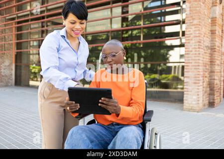 A professional Latin American woman assists a young African American woman in a wheelchair, using a digital tablet outdoors near a modern office build Stock Photo