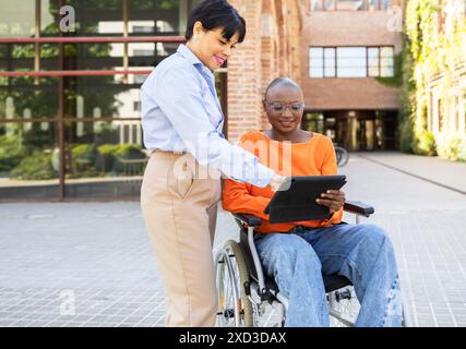 A professional black businesswoman assists her Latin American colleague, who is in a wheelchair, using a digital tablet outside an office building Stock Photo