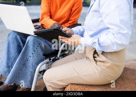 A wheelchair user and a colleague engage in a business discussion outdoors while using digital devices, highlighting inclusivity and teamwork Stock Photo