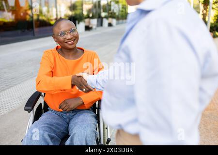 A smiling African American woman in a wheelchair wearing orange shakes hands with a woman in a blue shirt, symbolizing inclusivity and professionalism Stock Photo