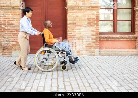 A Latin businesswoman pushes the wheelchair of an African American colleague, highlighting diversity and support in the workplace Stock Photo