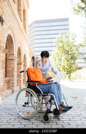 An image showcasing a young African American woman in a wheelchair and a Latin businesswoman using a laptop outdoors, embodying inclusivity and teamwo Stock Photo