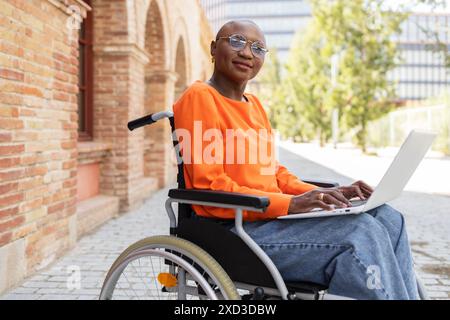 A confident young black woman in a wheelchair uses a laptop on a sunny path beside a historic brick building She dresses smartly in a bright orange to Stock Photo