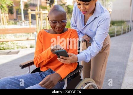 A latin businesswoman assists her black colleague, who is using a wheelchair, with a mobile phone while working outdoors Stock Photo