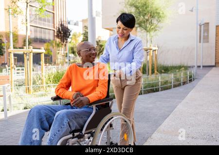 A supportive Latin businesswoman assists her African American colleague in a wheelchair, highlighting inclusion and teamwork in a sunny urban setting Stock Photo