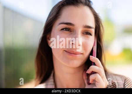 A young woman is engaged in a conversation on her mobile phone, looking away from with a thoughtful expression, outdoors during daylight. Stock Photo