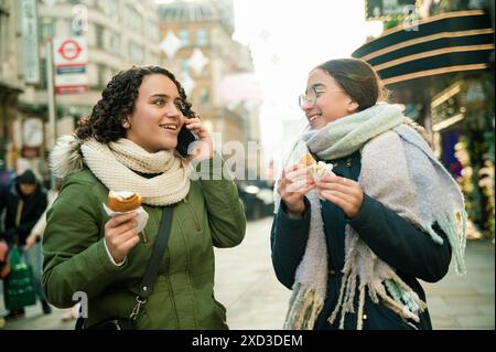 Two young women laugh and eat while one talks on the smartphone in a bustling London street. Stock Photo
