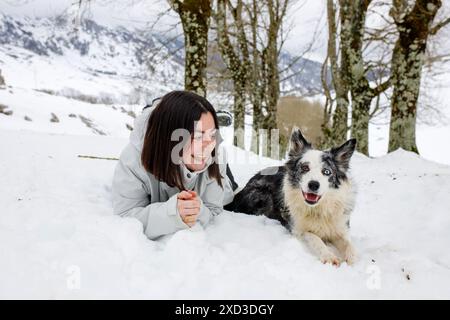 A woman and her Border Collie Blue Merle lie on the snow, both smiling and enjoying their time together in a snowy forest Stock Photo