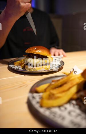 A person in a dark shirt is about to slice a juicy, stacked gourmet burger served on a plate with a side of golden fries at a modern eatery Stock Photo