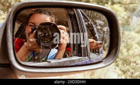 A woman takes a photo reflected in a vehicle's side mirror, with her Vizsla dog looking out from the passenger seat amidst a natural backdrop Stock Photo