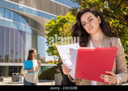 Two professional women engage in a discussion outside a modern office building One holds documents and a red folder, focusing intently Stock Photo