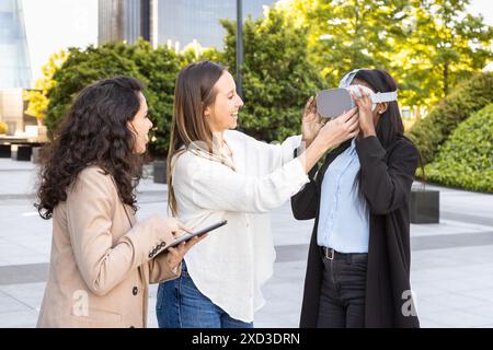 Three professional women of diverse backgrounds are collaborating outdoors, using a virtual reality headset in an urban park with modern buildings aro Stock Photo