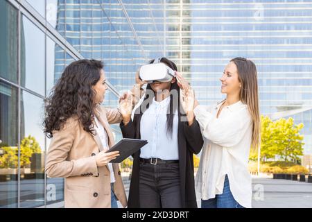 Three multiethnic professional women engage in testing virtual reality equipment outside a modern glass building, exploring advanced interactive techn Stock Photo