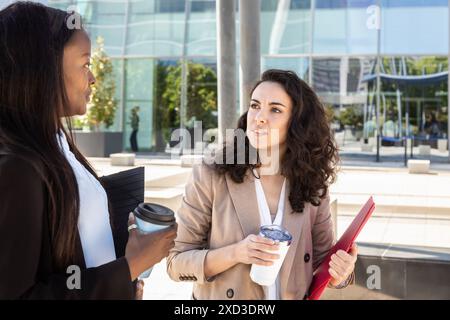 Two businesswomen in professional attire discuss work while holding coffee cups in a modern urban setting, reflecting active business communication an Stock Photo