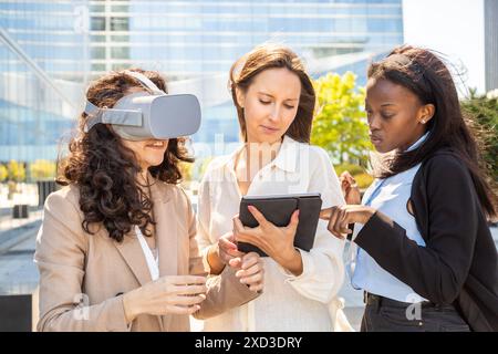 Three multiracial professional women engage with virtual reality technology outdoors, with one experiencing VR and the others managing the device Stock Photo