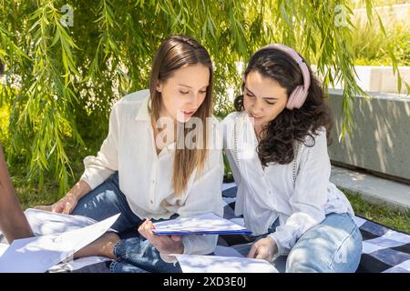 Two women, one wearing a white blouse and jeans, and the other a white tunic and headphones, are working together over papers and a clipboard on a pic Stock Photo