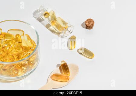 A well-lit, high-resolution image showcasing omega 3 capsules in a glass bowl, wooden spoon, and tipped glass vial on a white surface Stock Photo