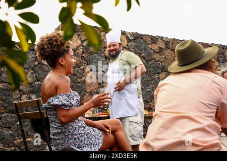 A joyful gathering of friends outdoors, featuring a Caucasian male chef in an apron cooking BBQ and a smiling African American woman enjoying a drink. Stock Photo