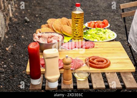 A ready-to-grill outdoor barbecue selection featuring raw meats, buns, and salads on a wooden board, complete with condiments and a bottle of oil. Stock Photo