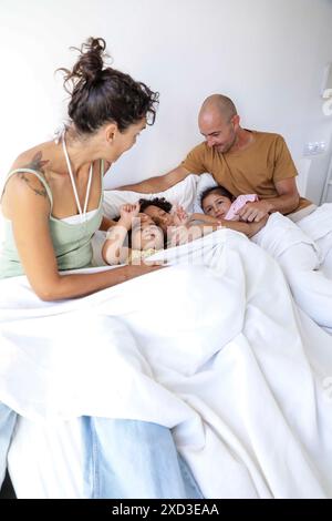 A multiethnic family enjoys a morning moment together in bed, two children wake up between their smiling parents, depicting a scene filled with warmth Stock Photo