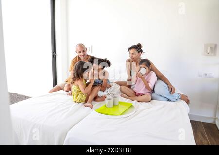 A multiethnic family shares a joyful morning in a sunlit bedroom, with parents and three children enjoying breakfast in bed, creating a warm, loving a Stock Photo