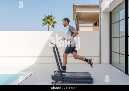 A focused young man jogs on a treadmill placed outdoors, enjoying a sunny day with palm trees and modern architecture in the background Stock Photo