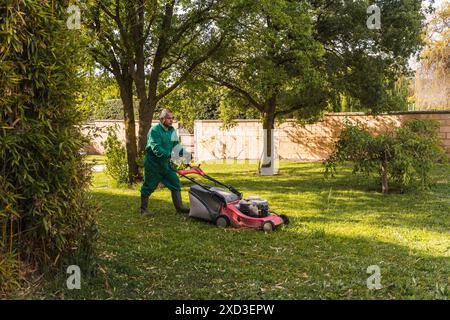 A middle-aged man in green work attire mows a lush garden with a modern lawn mower, surrounded by mature trees on a sunny day Stock Photo
