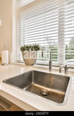 A kitchen sink in front of a window with blinds on the windowsilling it is white and there is a pot of flowers Stock Photo