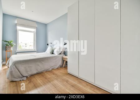 A contemporary bedroom featuring light blue walls, a large window, wooden flooring, and a minimalistic white wardrobe. Stock Photo