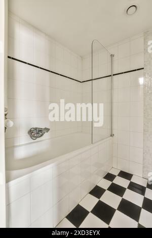 A modern bathroom featuring a white bathtub with a glass shower screen, black and white checkered floor, and white tiled walls with a black accent str Stock Photo